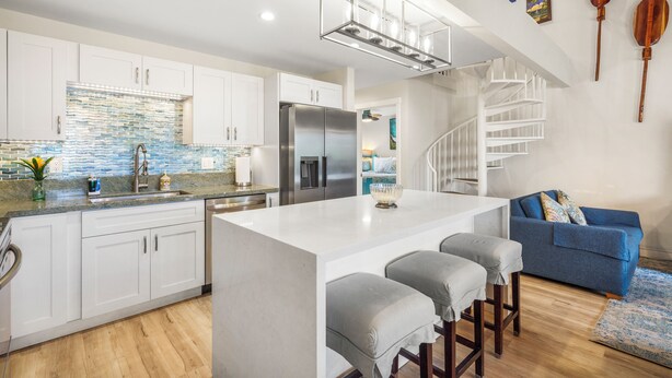 Una cocina moderna con armarios blancos, una isla central con taburetes de bar, un refrigerador de acero inoxidable y una lámpara colgante.