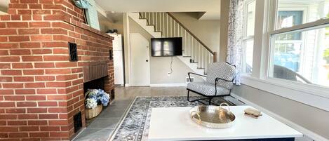 A living room with a brick fireplace, a patterned rug, a white table with a tray, and a chair.