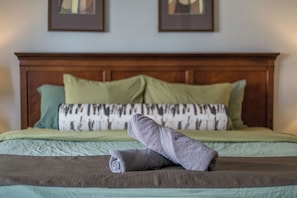 Una cama bien hecha con un edredón verde, una almohada estampada y una toalla enrollada.