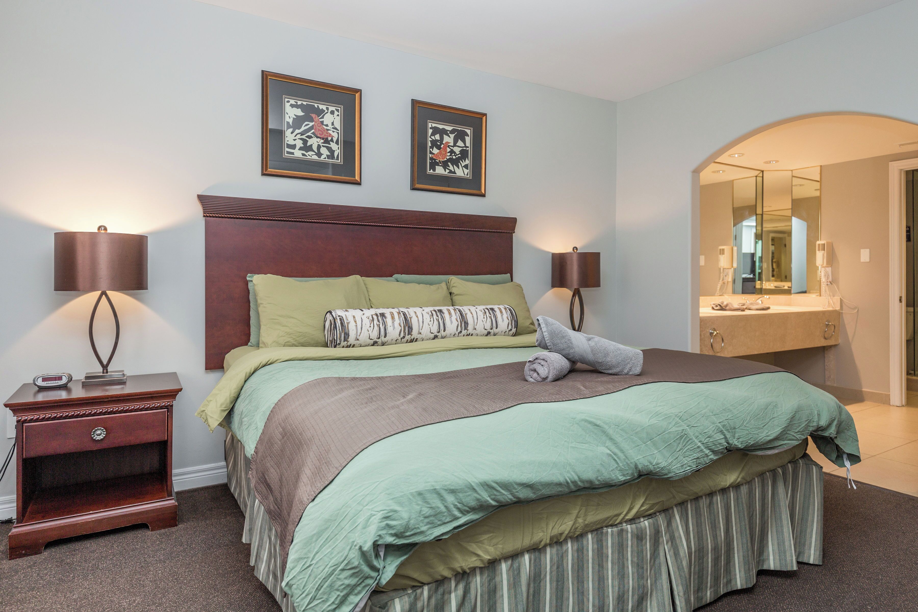 A bedroom with a large bed, two bedside tables, a nightstand, a mirror, and framed artwork on the wall.