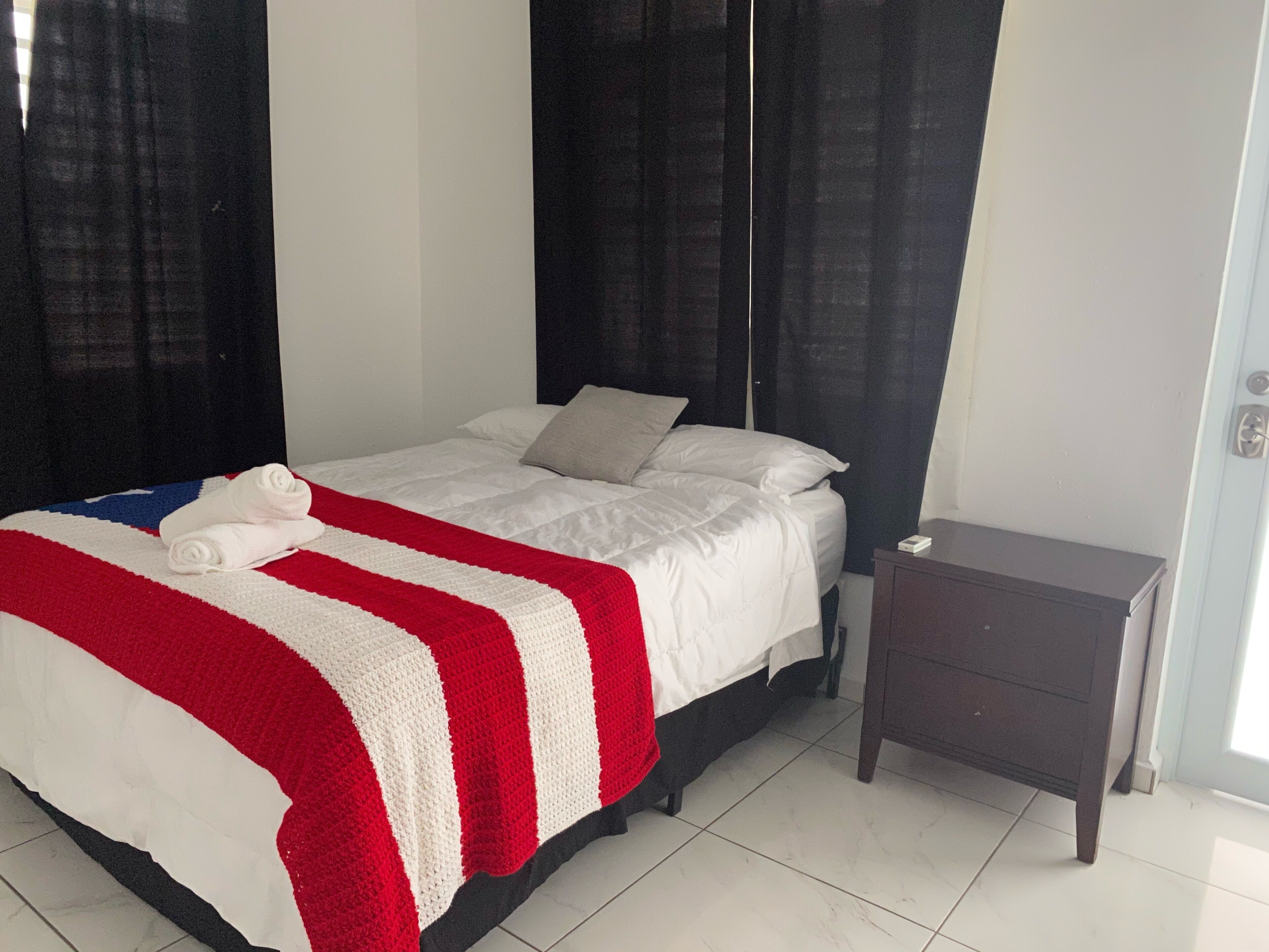 Una cama bien hecha con ropa blanca y una colcha a rayas rojas y blancas, una pequeña mesita de noche y una cortina oscura.