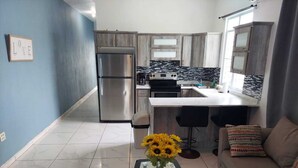 A modern kitchen with a stainless steel refrigerator, a gas stove, and a built-in oven. There is a dining area with a glass table and two black bar stools. - Jobos Luxury Condos (Isabela)