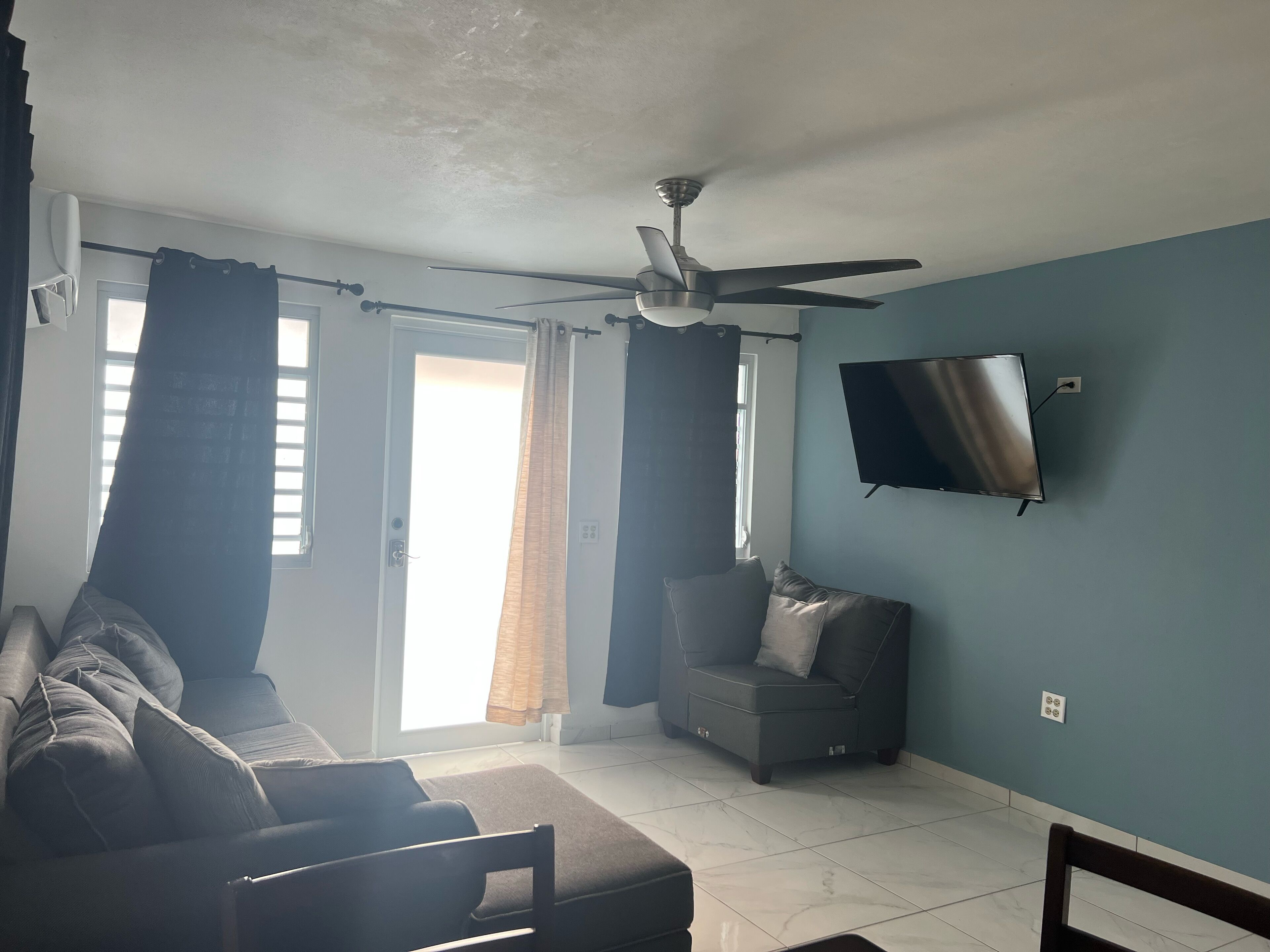 A living room with a sofa, a chair, a television mounted on the wall, a ceiling fan, and a window with curtains.