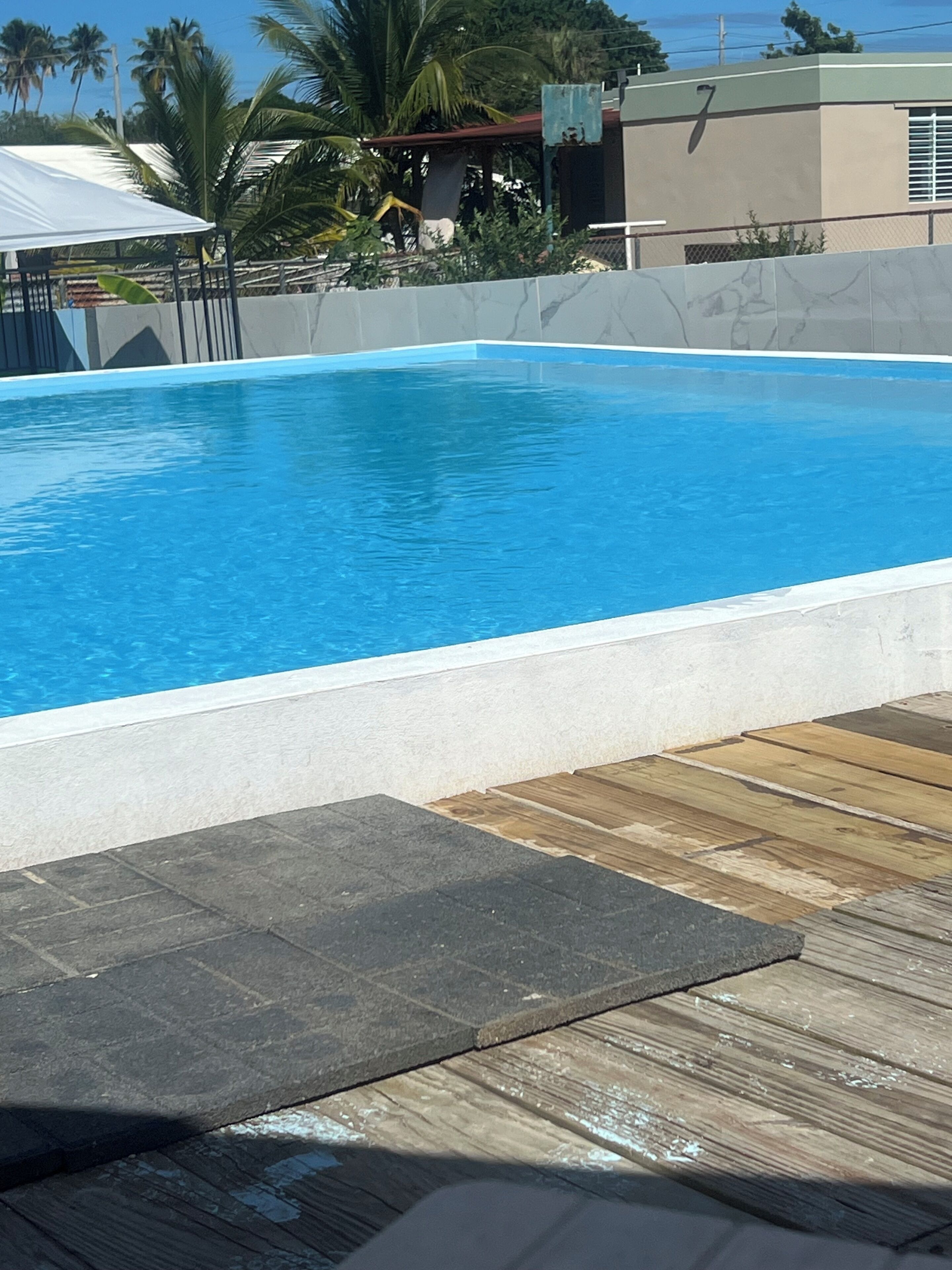 Une piscine avec une terrasse en bois au premier plan, entourée de palmiers et d’un bâtiment portant le chiffre « 7 ».