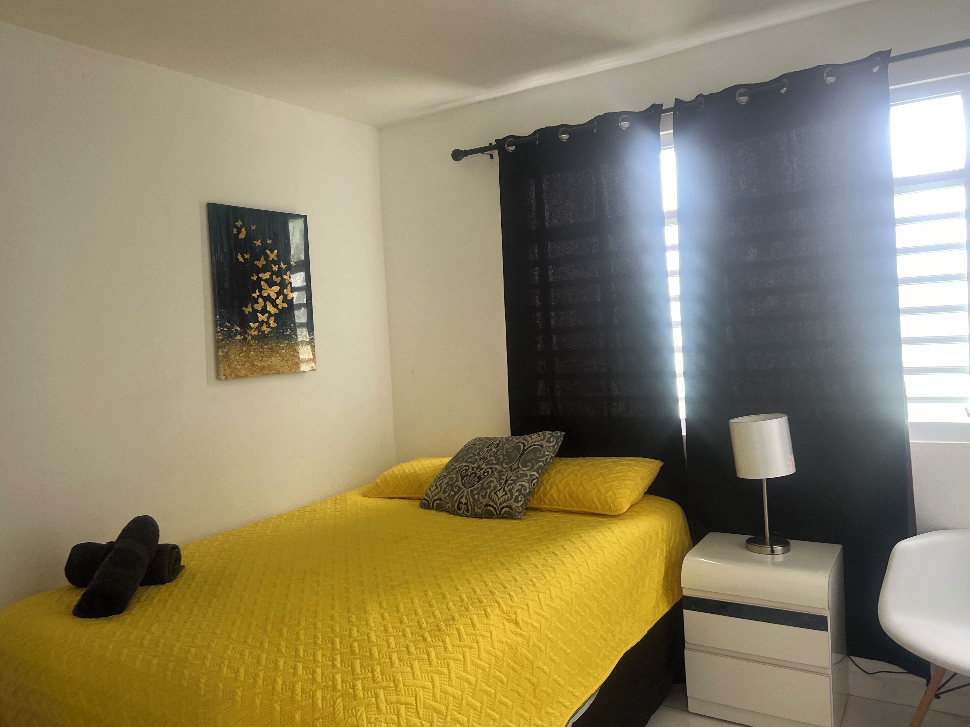 Una camera da letto con un letto giallo, una tenda nera, un comodino bianco, una lampada e un quadro appeso al muro.
