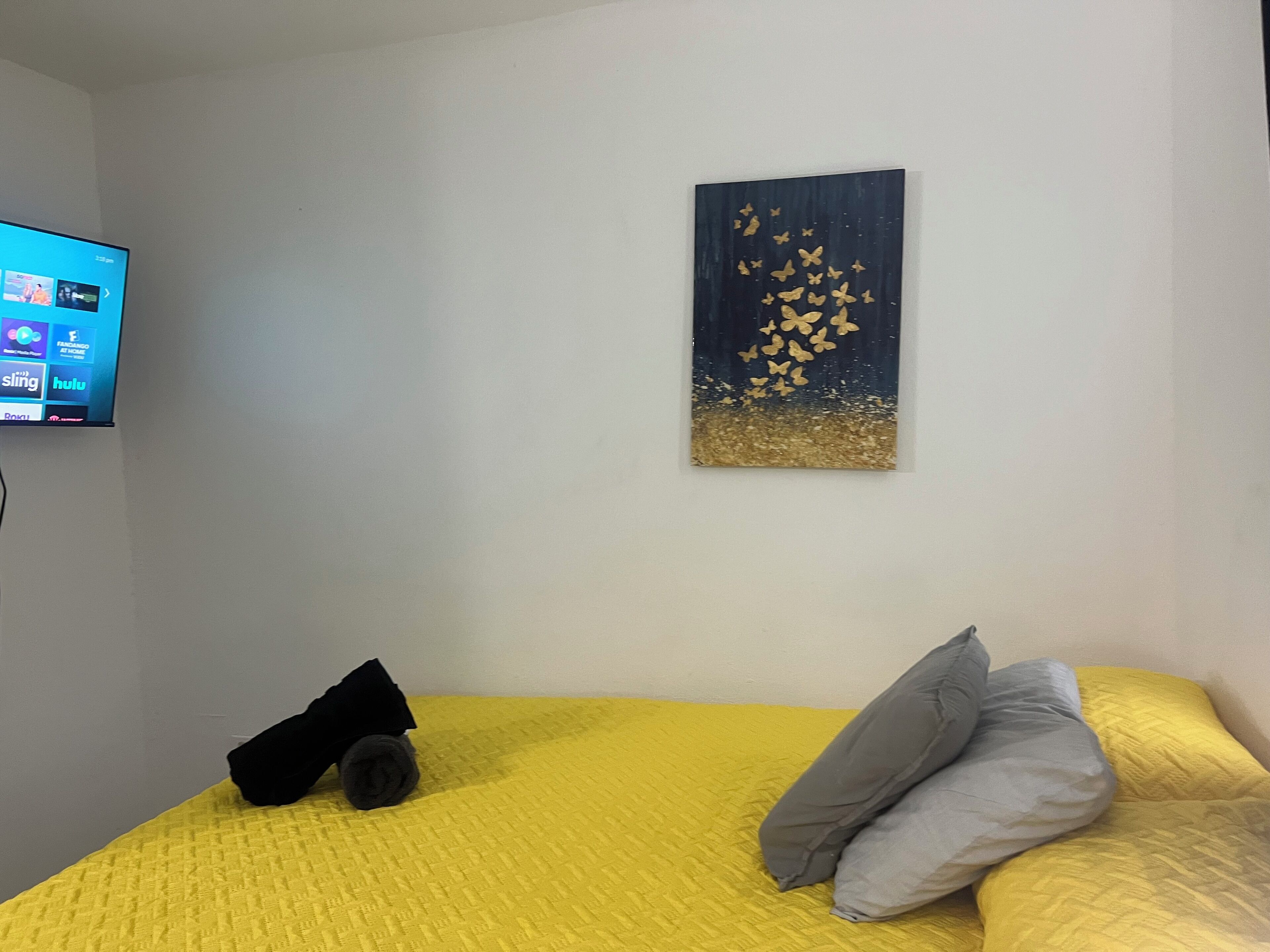 Ein Zimmer mit einem gelben Bett, einem grauen Kopfkissen mit schwarzem Muster und einem Schmetterlingsbild an der Wand.