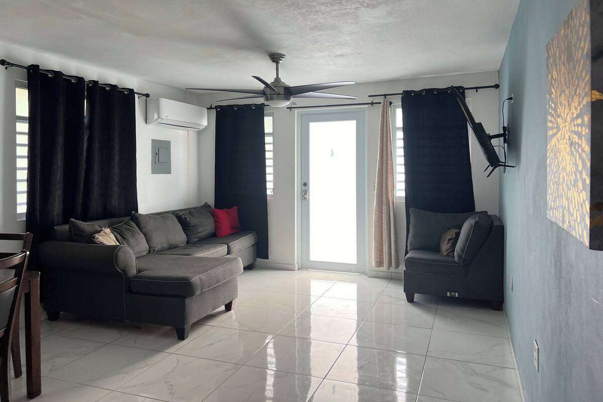 A living room with a grey sofa, a ceiling fan, and a door leading outside.
