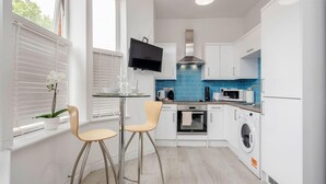 Une cuisine compacte avec des meubles blancs, un pare-feu bleu, un four encastré, un four à micro-ondes et un lave-linge. On y trouve deux tabourets et une petite table sur laquelle est posée une orchidée en pot.