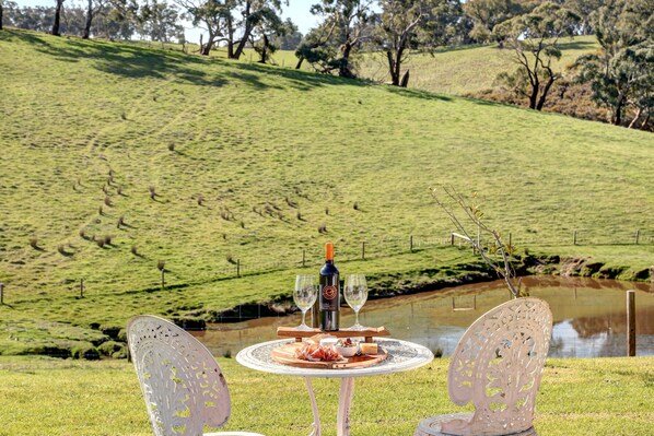 A table with a bottle of wine, two glasses, and a plate of food set on a grassy hill overlooking a body of water. - Meadows Farmhouse - Adelaide Hills (Meadows)