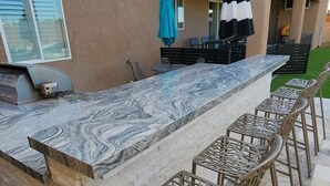 Cozinha externa com bancada de mármore, quatro banquetas e churrasqueira.