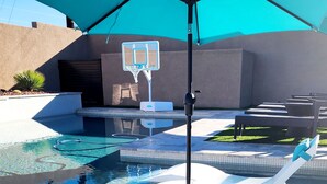 Uma área de piscina com cesta de basquete, espreguiçadeiras e um grande guarda-sol.