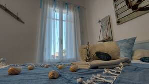 Ein Schlafzimmer mit Bett, einem Fenster mit durchsichtigen Vorhängen, einer Wandregal mit Dekoration und einem blauen, strukturierten Teppich.