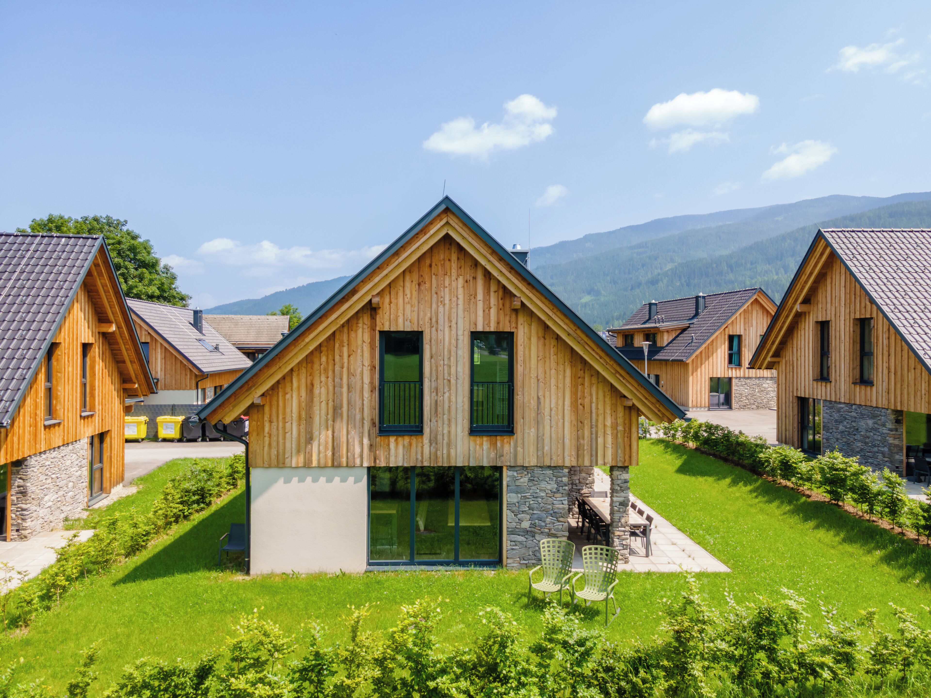 Un chalet en bois, doté de fondations en pierre, entouré d’une végétation luxuriante et situé au pied d’une chaîne de montagnes.
