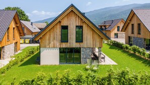 Een houten chalet met een stenen fundering, omringd door weelderige begroeiing en met een bergachtig landschap op de achtergrond.