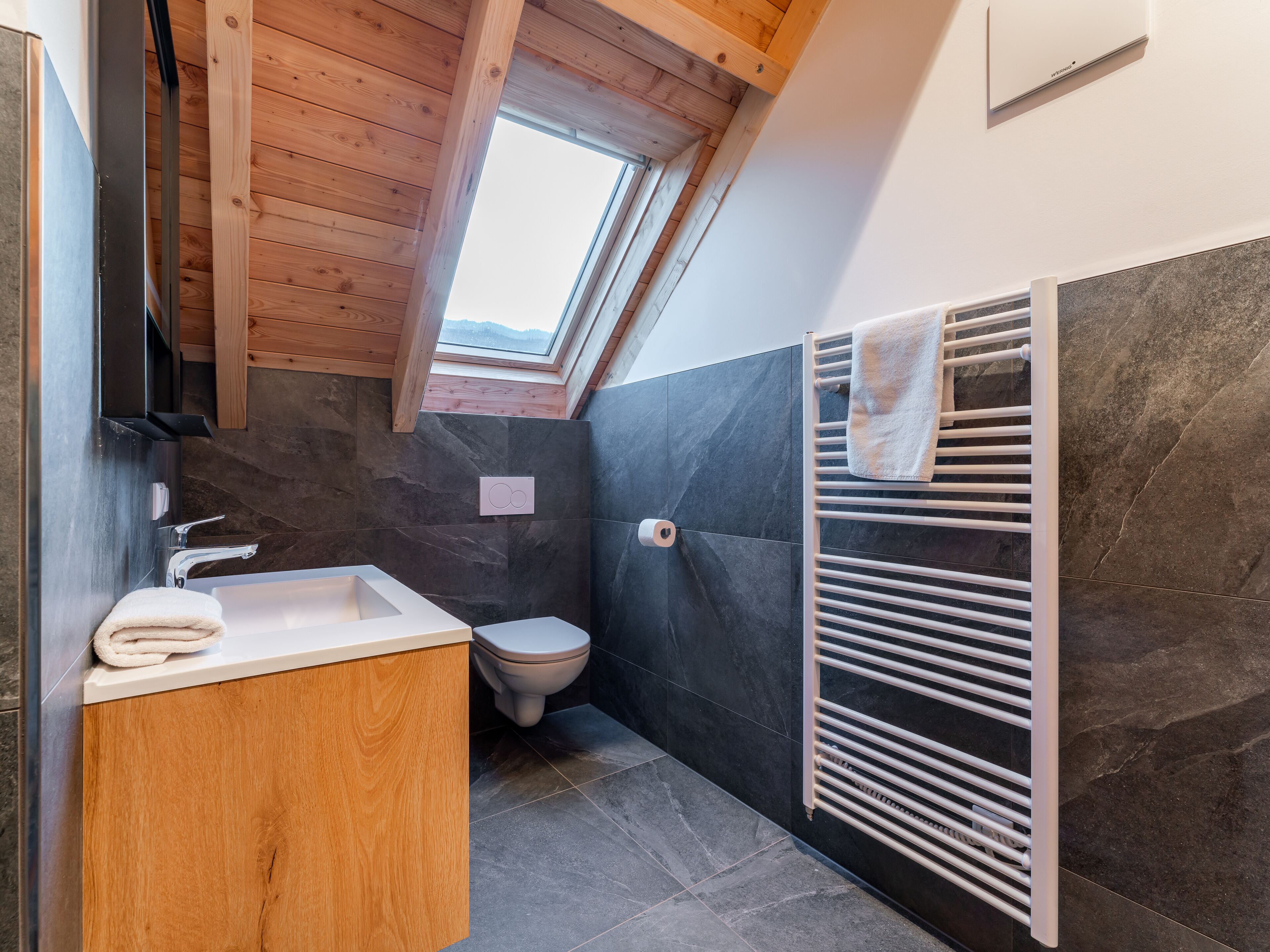 Un baño moderno con techo de madera, tragaluz, lavamanos blanco, inodoro y toallero.