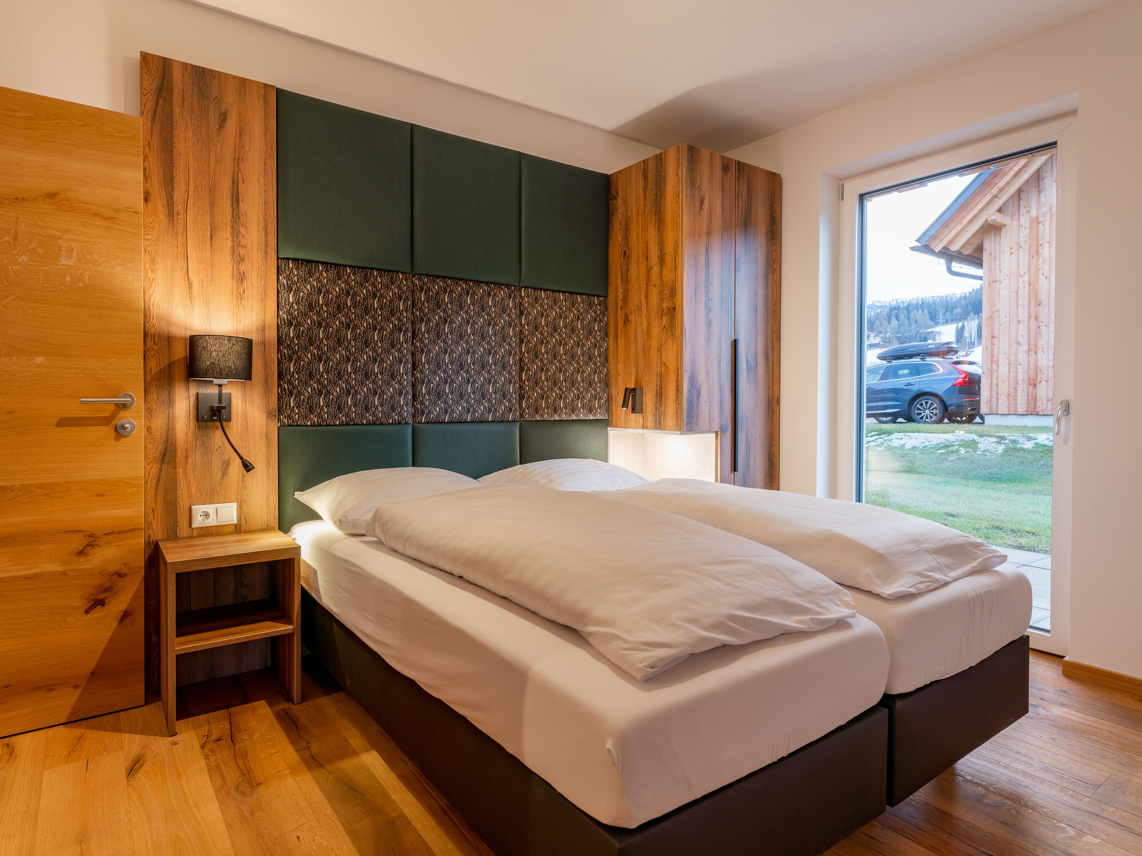 A modern bedroom with a wooden wardrobe, a bedside table, a headboard with a textured panel, and a view of a car and building outside.