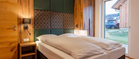 A modern bedroom with a wooden wardrobe, a bedside table, a headboard with a textured panel, and a view of a car and building outside.
