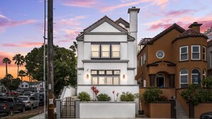 A two-story house with a white facade and brown trim, featuring a large window and a small porch area with plants. - Pet-friendly! Near downtown Oceanside - Sea Spray (Oceanside)