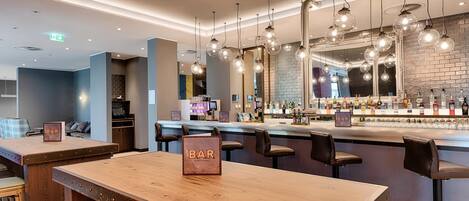Zona de bar con mesas y taburetes de madera, una barra con diferentes botellas e iluminación colgante.