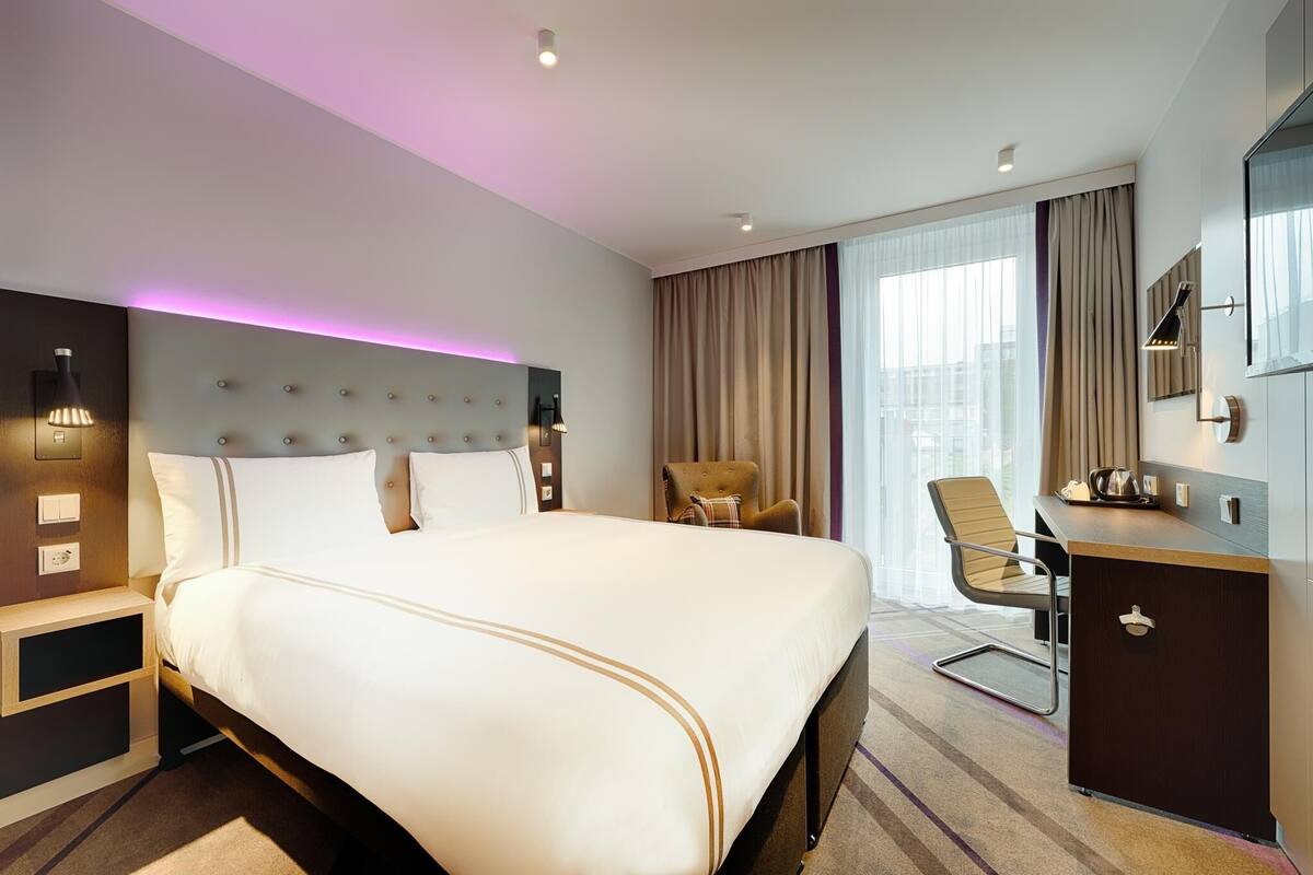 A hotel room with a large bed, a desk, a chair, and a view of the city through the window.