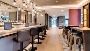 A bar area with multiple stools, a counter with bar signs, hanging light fixtures, and a large mirror. - Premier Inn Hamburg City Klostertor (Hamburg)