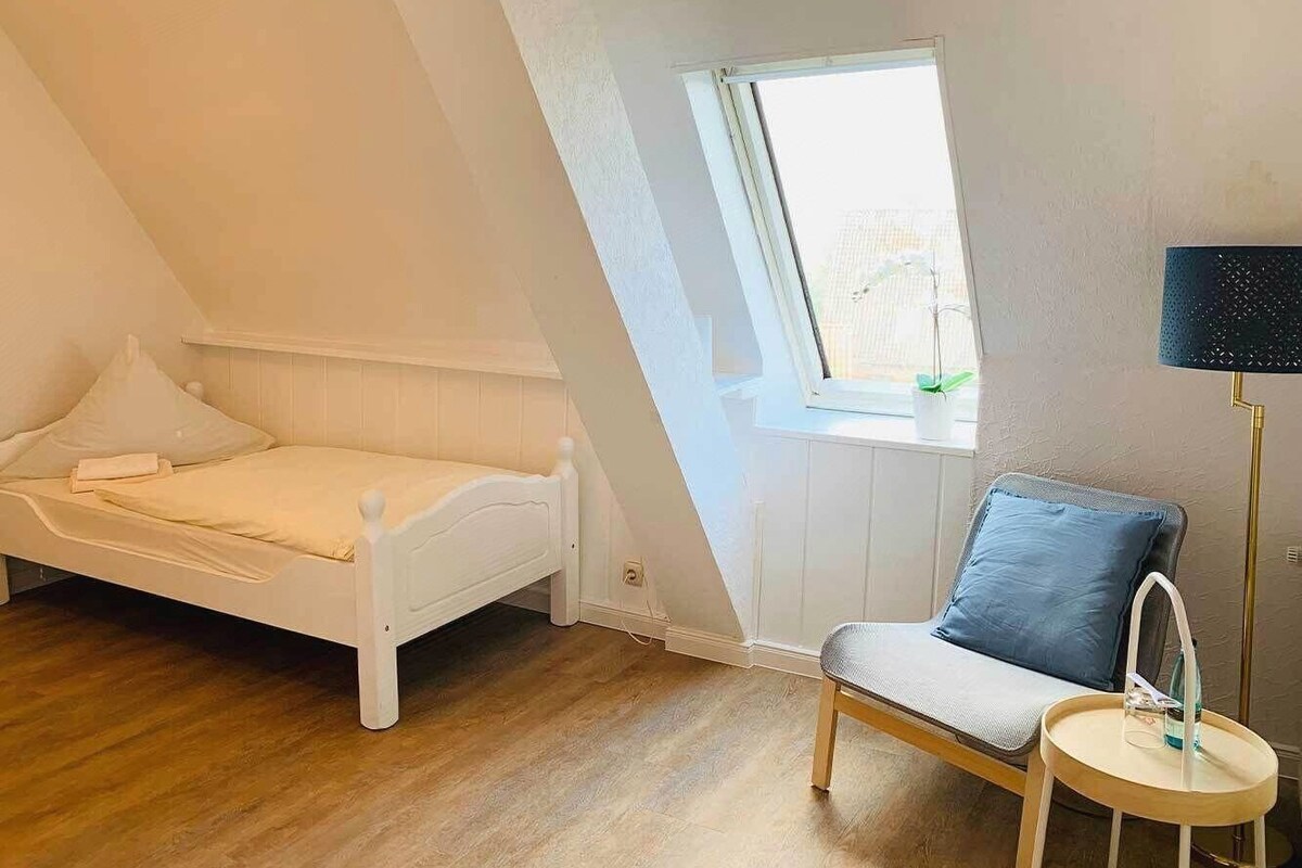 A bedroom with a sloped ceiling, a bed, a chair, a small table, and a window.