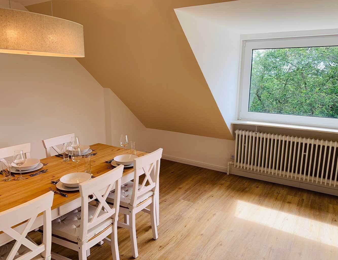Comedor con una mesa de madera dispuesta para cuatro, sillas blancas, un radiador blanco y una ventana con vistas a árboles.
