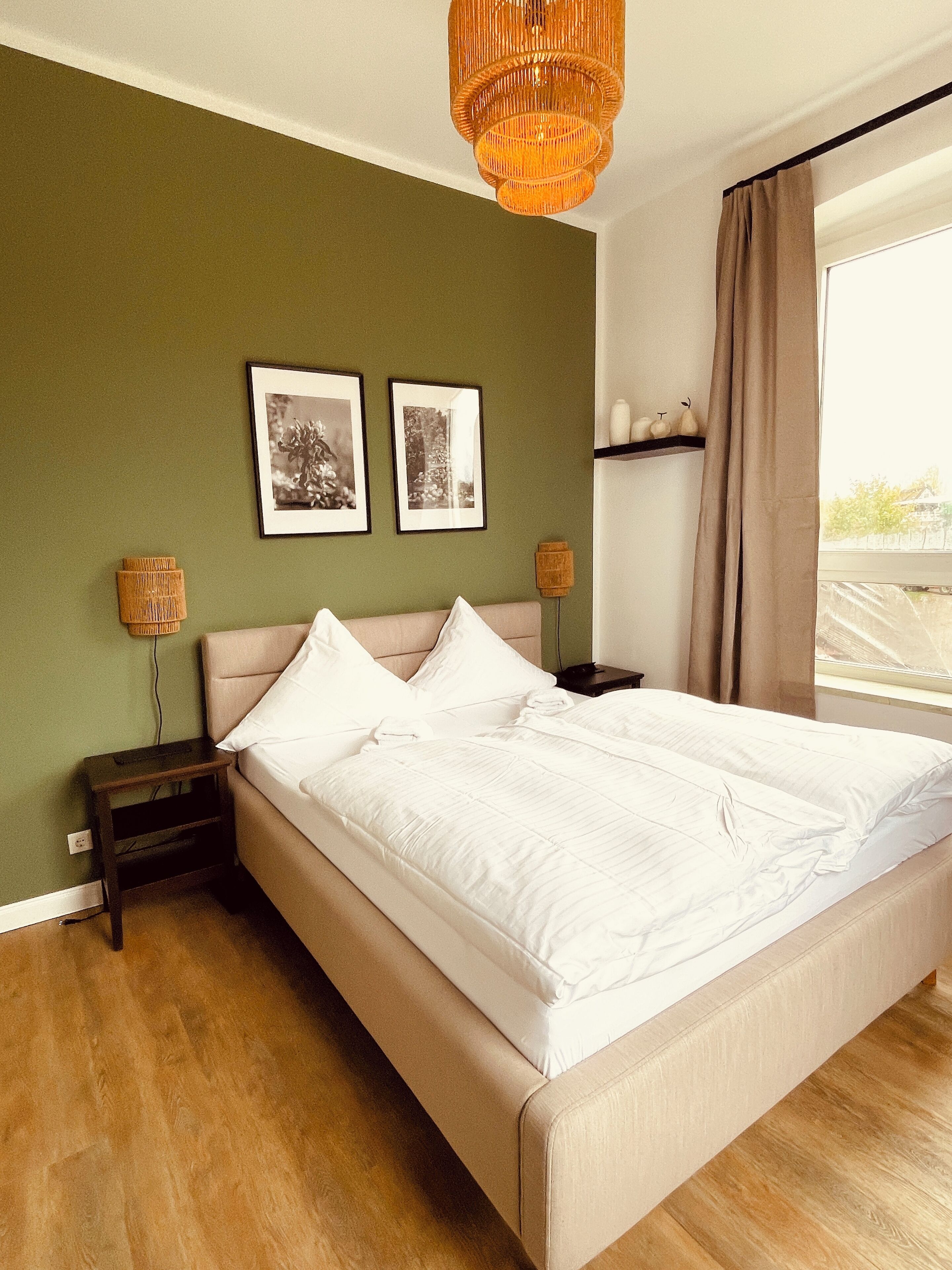 Une chambre à coucher avec un grand lit, deux tableaux encadrés au mur, une table de chevet et une fenêtre avec des rideaux.