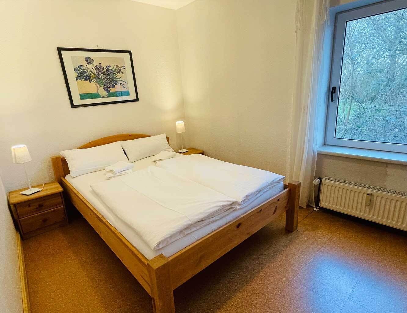 Cama bem arrumada em quarto com janela, mesa de cabeceira e quadro na parede.