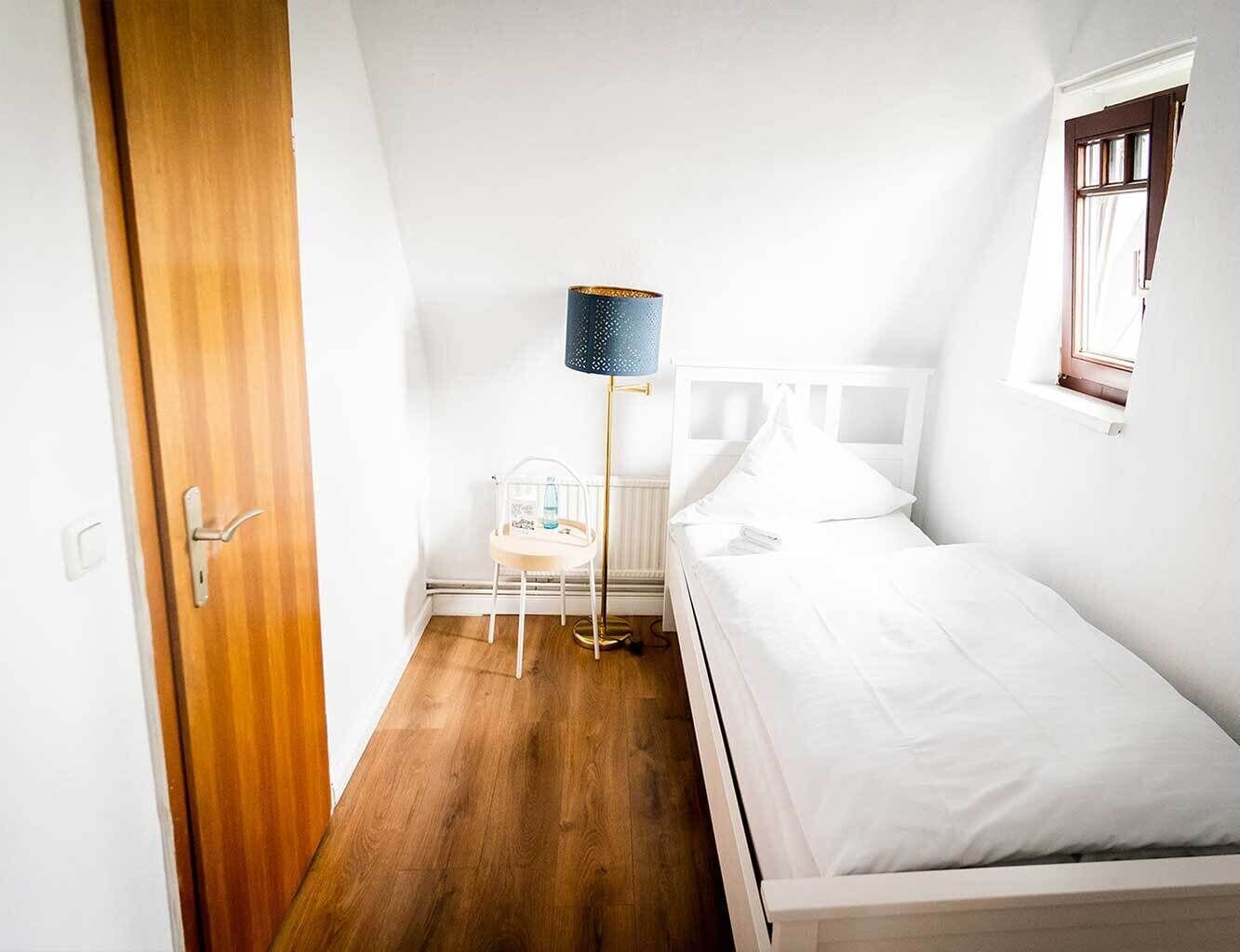 Un dormitorio con piso de madera, una cama con ropa de cama blanca, una mesita de noche con una lámpara y una ventana con cortinas.