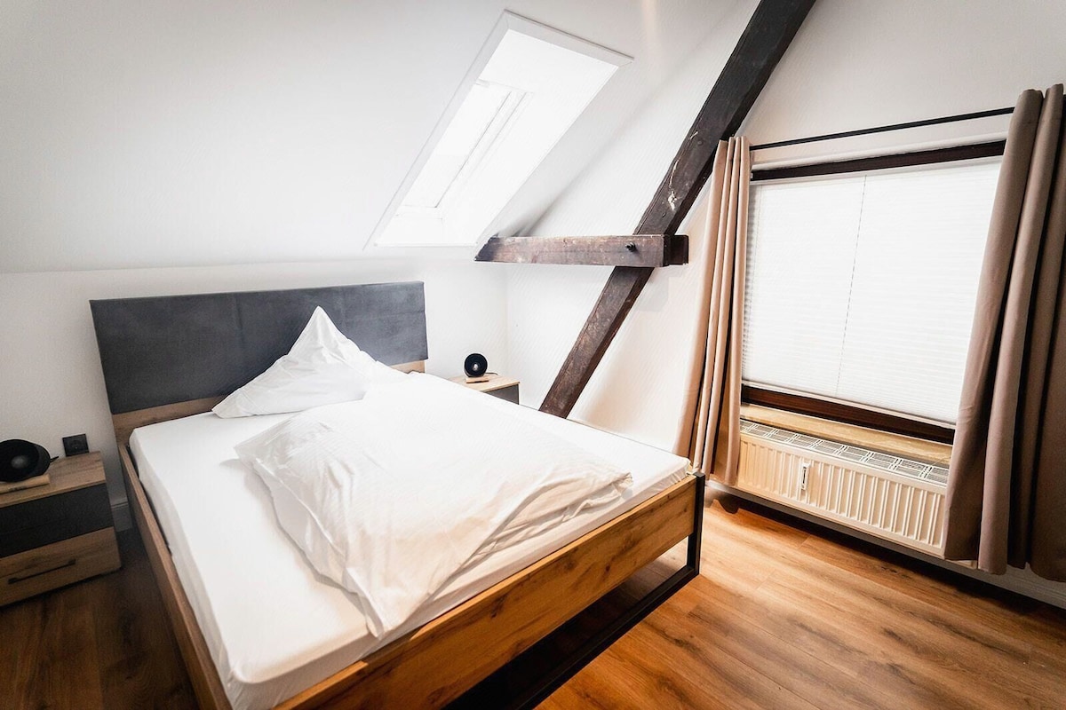 A modern bedroom with a sloped ceiling, a wooden bed frame, a bedside table with a lamp, and a window with curtains.