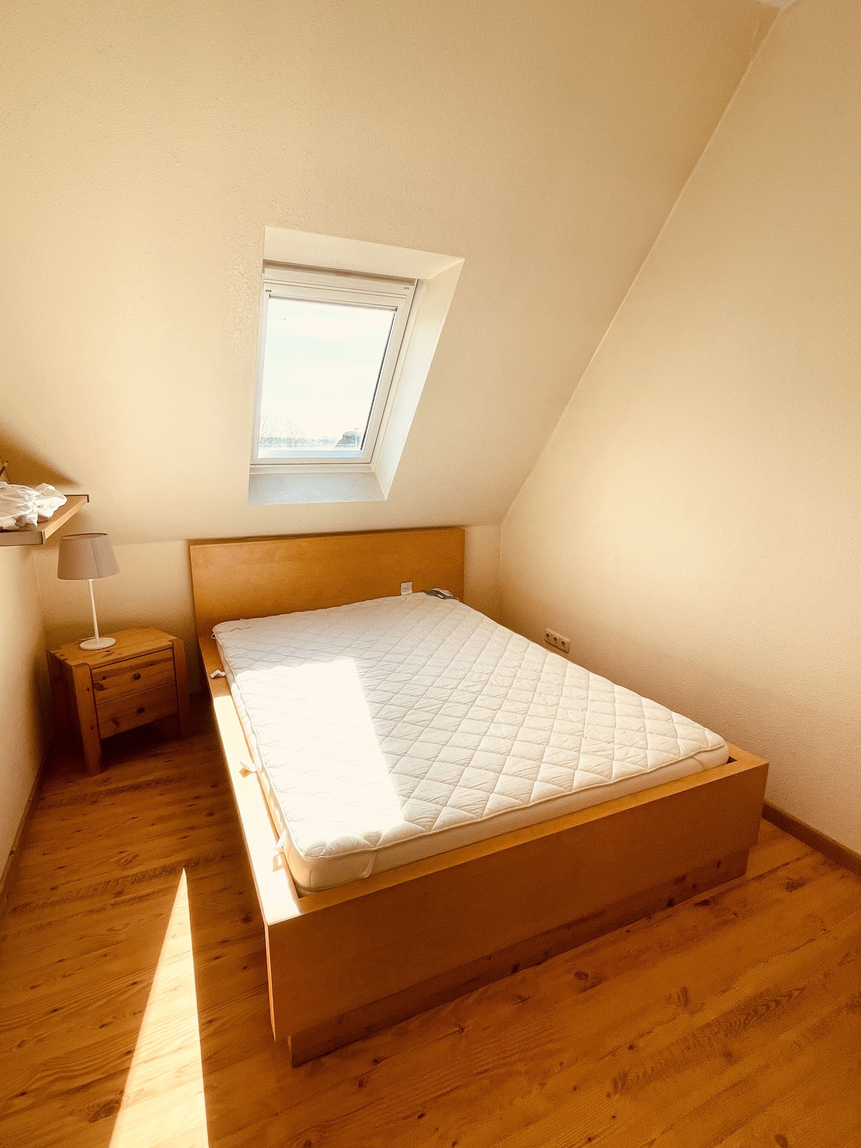 Ein kleiner, gut beleuchteter Raum mit schrägem Dach, einem Einzelbett, einem Nachttisch, einer Lampe und einem Fenster.