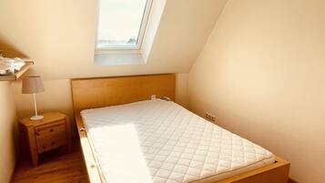 Ein kleiner, gut beleuchteter Raum mit schrägem Dach, einem Einzelbett, einem Nachttisch, einer Lampe und einem Fenster.