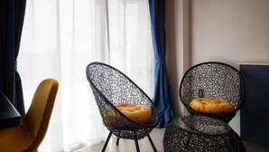 Zwei schwarze Stühle mit Drahtgestell und orangefarbenen Kissen, ein gelber Stuhl und ein Glastisch in einem Raum mit großem Fenster und durchsichtigen Vorhängen.