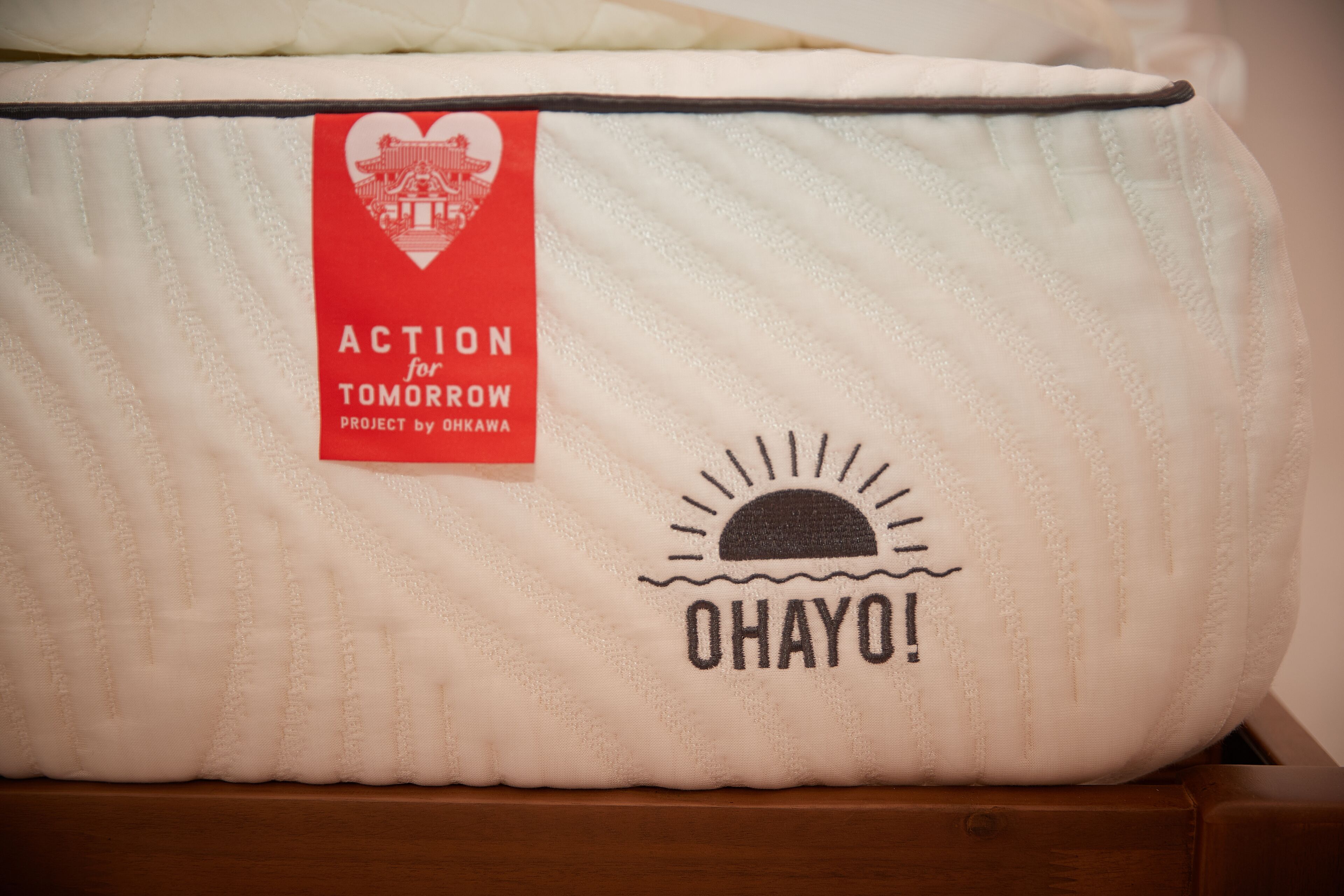 A white quilted bag with a red label that reads "ACTION for TOMORROW PROJECT by OHKAWA" and a black sun design with the word "OHAYO!"