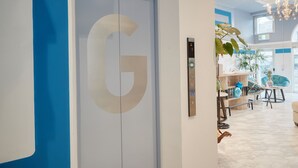 Ein modernes Interieur mit einem großen 'G'-Logo an einer Tür, einer Chandelier und einer Sitzecke mit Pflanzen und einem blauen Sessel.