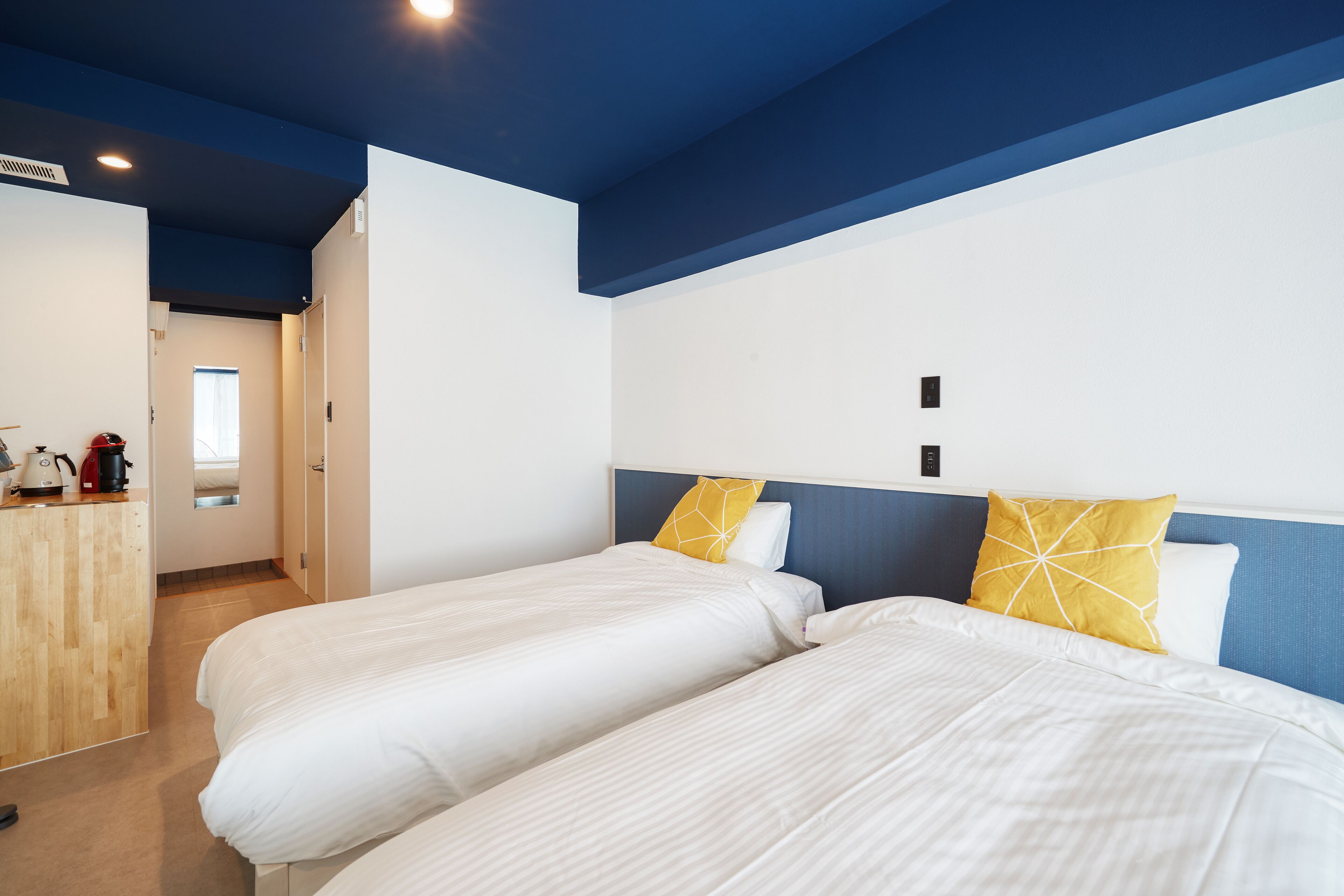A hotel room with two beds, a wooden desk, and a view of the hallway.
