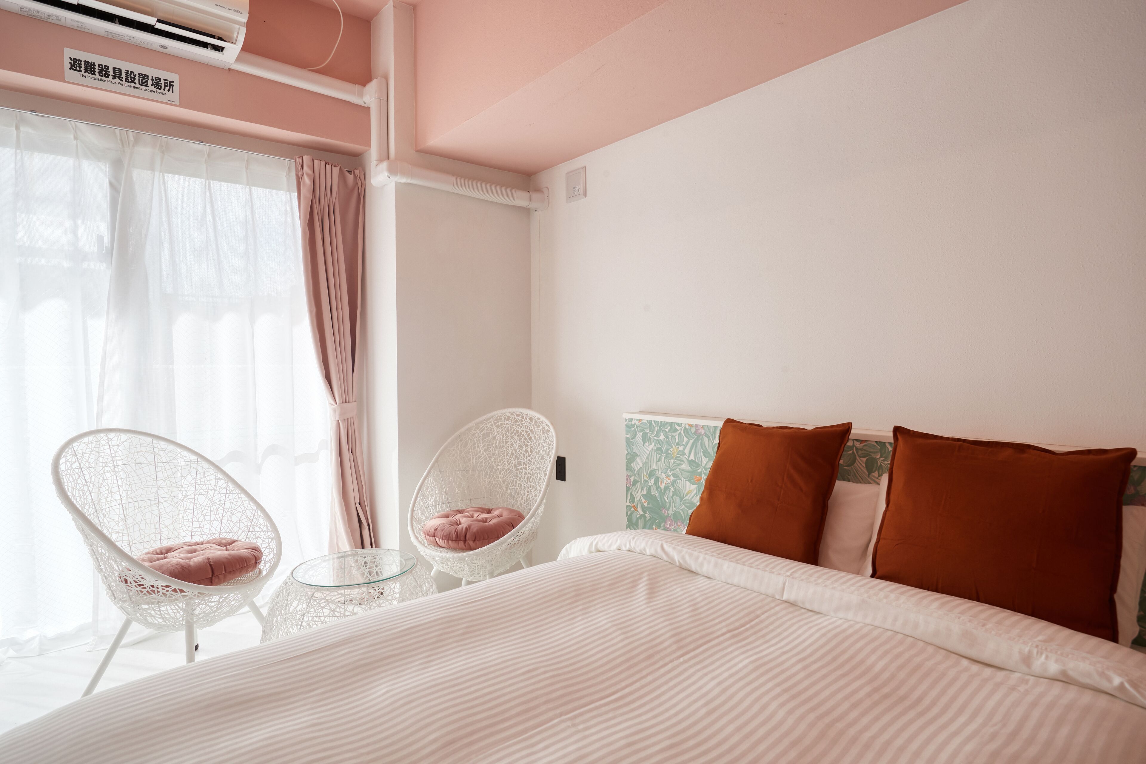 A hotel room with a bed, two chairs, a window with curtains, and an air conditioning unit.