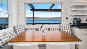 A dining area with a wooden table and white chairs, a vase with flowers, and a view of the sea through a large window.