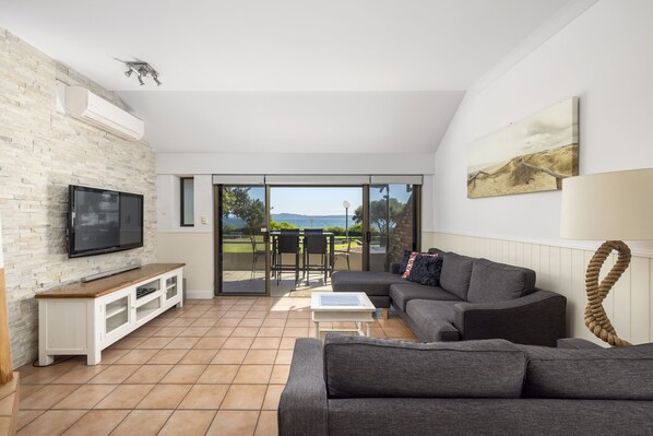 A modern living room with a grey sofa, a flat-screen TV, and a large window offering a view of a grassy area.