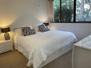 A neatly made bed with white bedding and patterned pillows, a bedside table with a lamp, and a window with a view of trees. - *Direct Access to Orion Beach* Jervis Bay Beachfront (Vincentia)