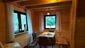 Une cabane en bois chaleureuse, dotée d’un coin repas avec une table et des chaises, de grandes fenêtres avec des rideaux et un luminaire suspendu.