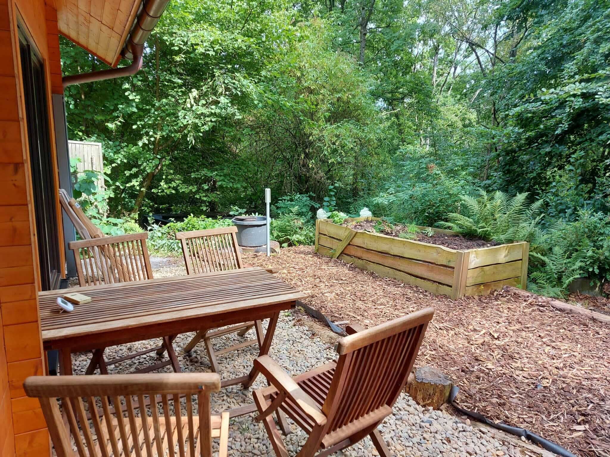Une terrasse extérieure avec une table et des chaises en bois, entourée d’un jardin agrémenté d’un parterre surélevé et d’un feuillage dense.