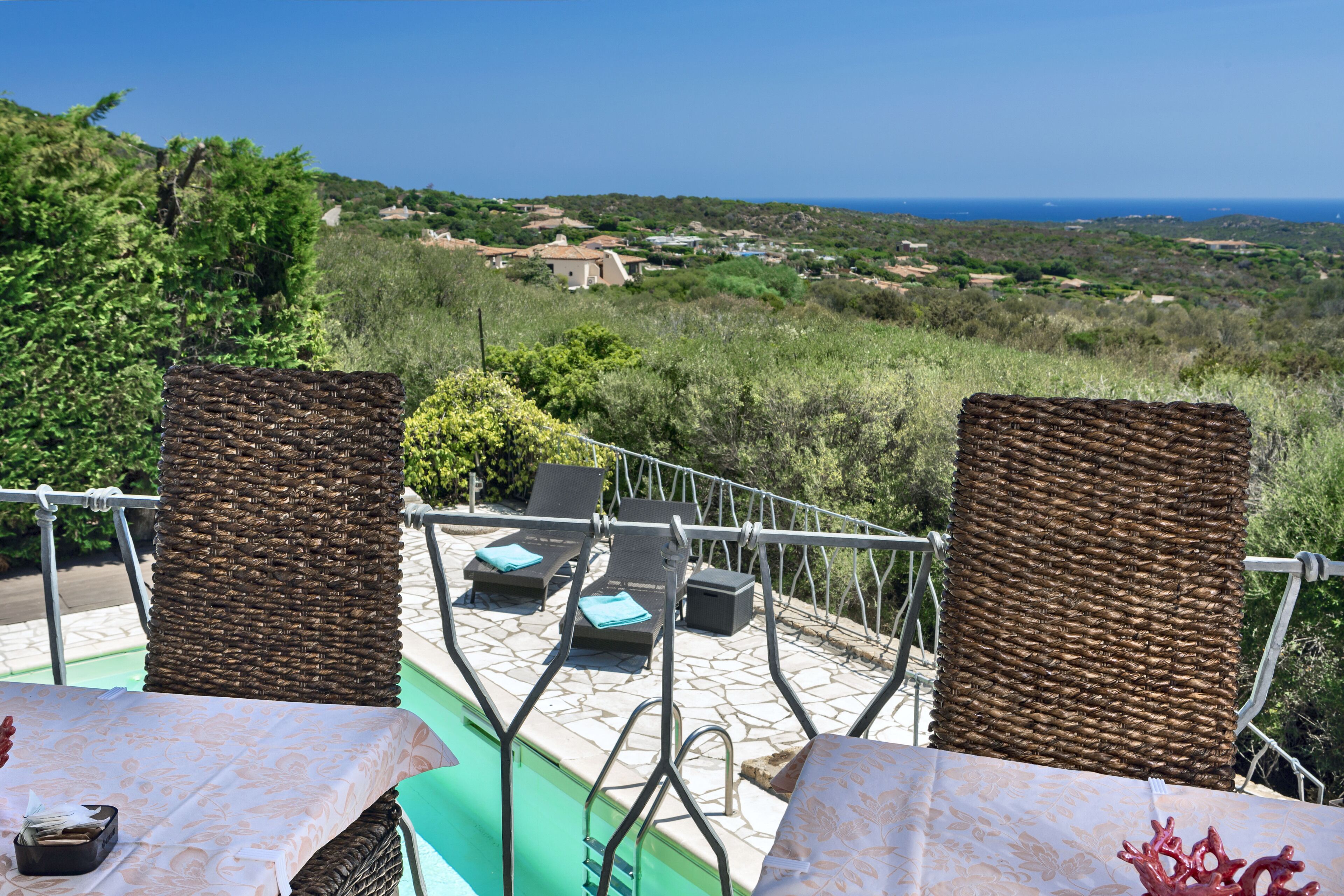 Une terrasse dotée de chaises et d’une table en osier, offrant une vue panoramique sur un paysage magnifique, avec la mer et des bâtiments éloignés à l’horizon.