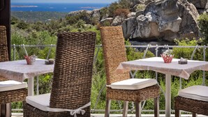 Mobiliario de mimbre para exteriores con cojines blancos y una mesa, sobre un fondo pintoresco de terreno rocoso y una vista distante del mar.
