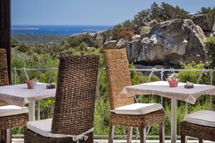 Mobiliario de mimbre para exteriores con cojines blancos y una mesa, sobre un fondo pintoresco de terreno rocoso y una vista distante del mar.
