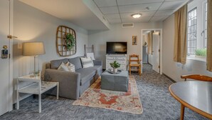 Una sala de estar con un sofá gris, una mesita auxiliar blanca, un reposapiés gris, un tapete estampado, un televisor y una silla de madera.