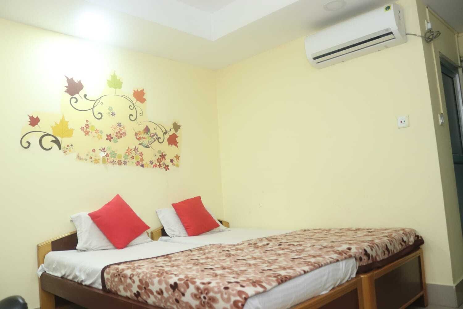A bedroom with a bed, two red pillows, a floral bedspread, and a wall decoration with autumn leaves and flowers.