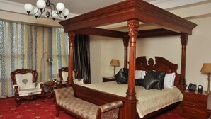 Un lit à baldaquin avec une structure en bois, un sol recouvert d’une moquette rouge, un banc recouvert d’un tissu blanc, une petite table ronde avec une lampe, et une table de chevet avec une lampe et un téléphone.