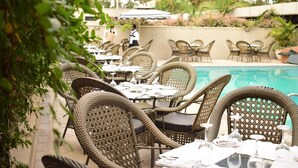Un espace extérieur aménagé pour les repas, avec des chaises et des tables en osier, une piscine et un serveur en arrière-plan.